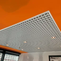 Sufit podwieszany rastrowy z wbudowanym oświetleniem punktowym na tle pomarańczowego stropu, widoczne okna i fragment ściany w stanie surowym.