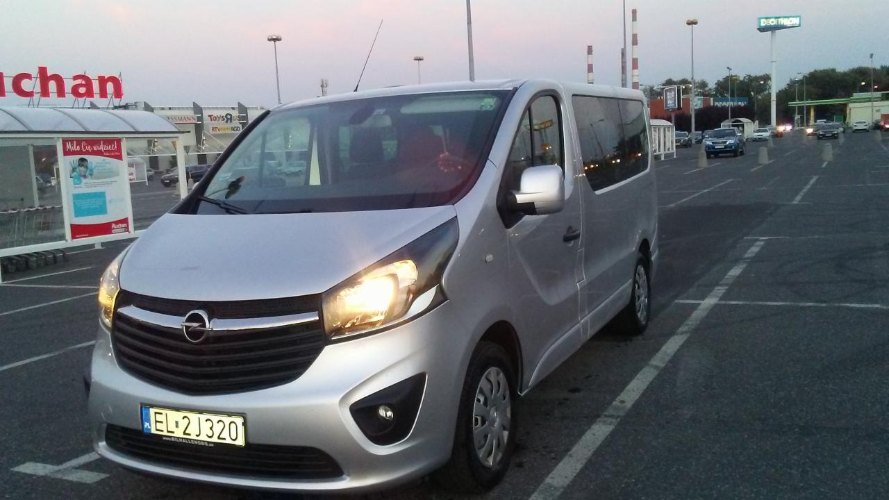 Srebrny Opel Vivaro zaparkowany na parkingu przed sklepem Auchan, z włączonymi światłami mijania.