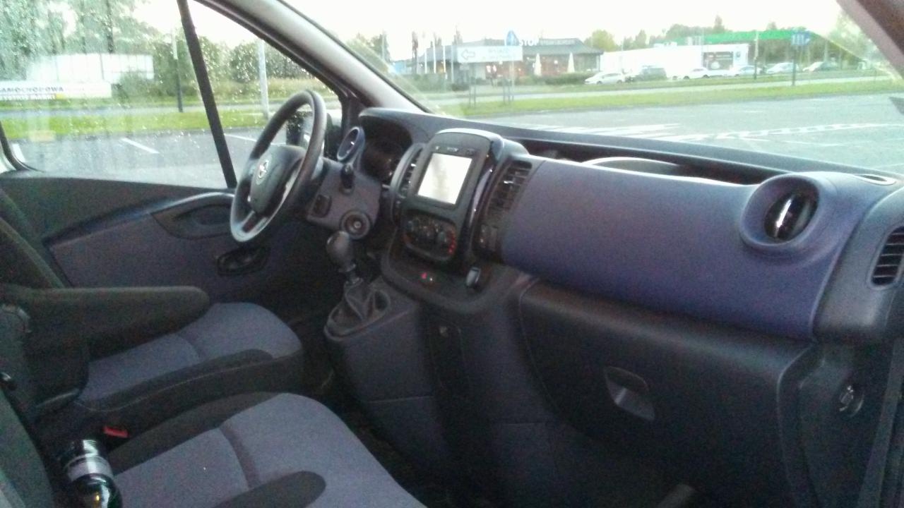 Wnętrze busa Opel Vivaro z widoczną deską rozdzielczą, kierownicą i fotelami w odcieniach szarości i granatu, z butelką wody w uchwycie na fotelu pasażera.