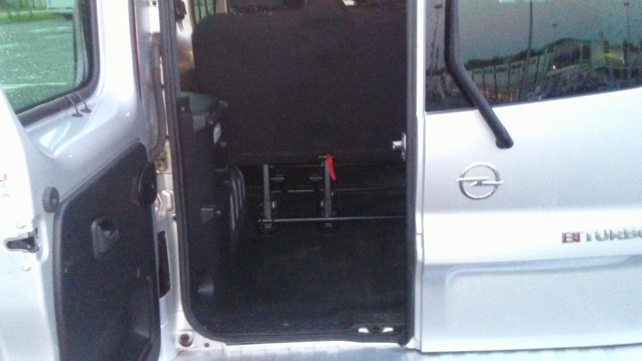 Wnętrze srebrnego Opla Vivaro z otwartymi drzwiami, widoczne zamontowane szyny do mocowania foteli lub wózków inwalidzkich.