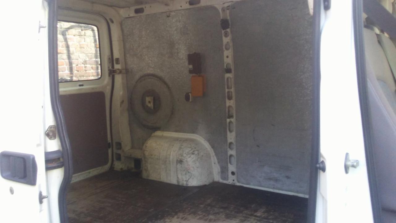 Wnętrze brudnego, białego busa transportowego z otwartymi drzwiami, widoczny szary panel boczny i podłoga z plamami.