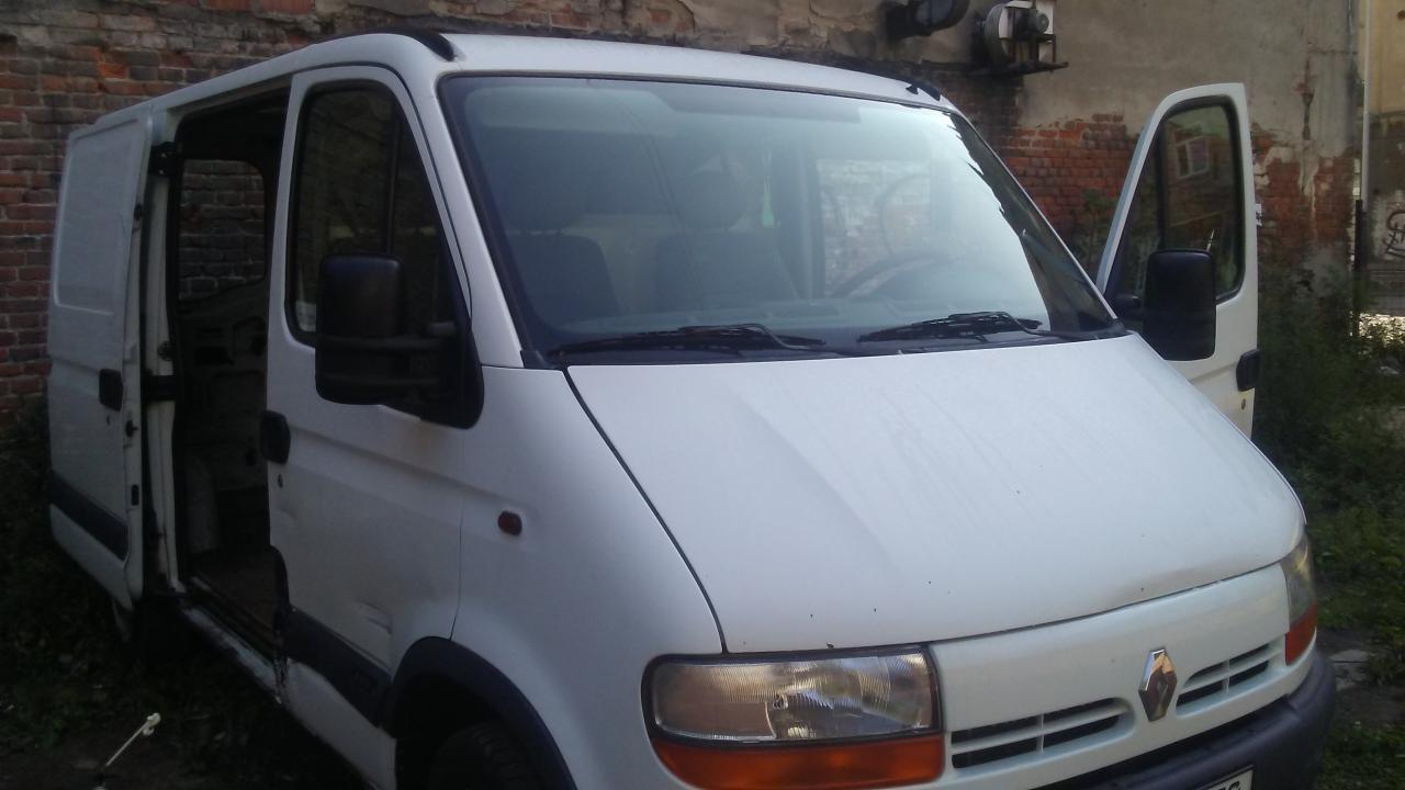 Biały bus Renault Master z otwartymi drzwiami bocznymi i kierowcy, zaparkowany na tle ceglanego muru z widocznymi śladami graffiti i roślinnością.