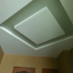 Sufit z podwieszanym, kwadratowym panelem centralnym, obniżonym i obramowanym zielonym pasem, widok z dołu na tle ścian pokrytych panelami i dwoma drzwiami.