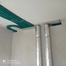 Szereg zielonych, karbowanych rur wentylacyjnych przymocowanych do sufitu i schowanych za szarą listwą, z dwoma pionowymi, srebrnymi rurami owiniętymi folią aluminiową, widocznymi na tle surowej...