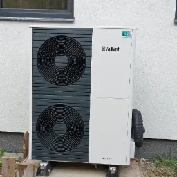 Biała pompa ciepła Vaillant aroTHERM zamontowana na betonowym fundamencie obok budynku z widocznym oknem.