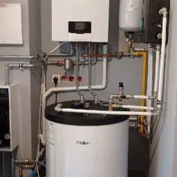 Biały, wiszący piec gazowy marki Vaillant z podłączonym zasobnikiem ciepłej wody, rurami miedzianymi i żółtą rurą gazową na tle szarej ściany i płytek podłogowych.