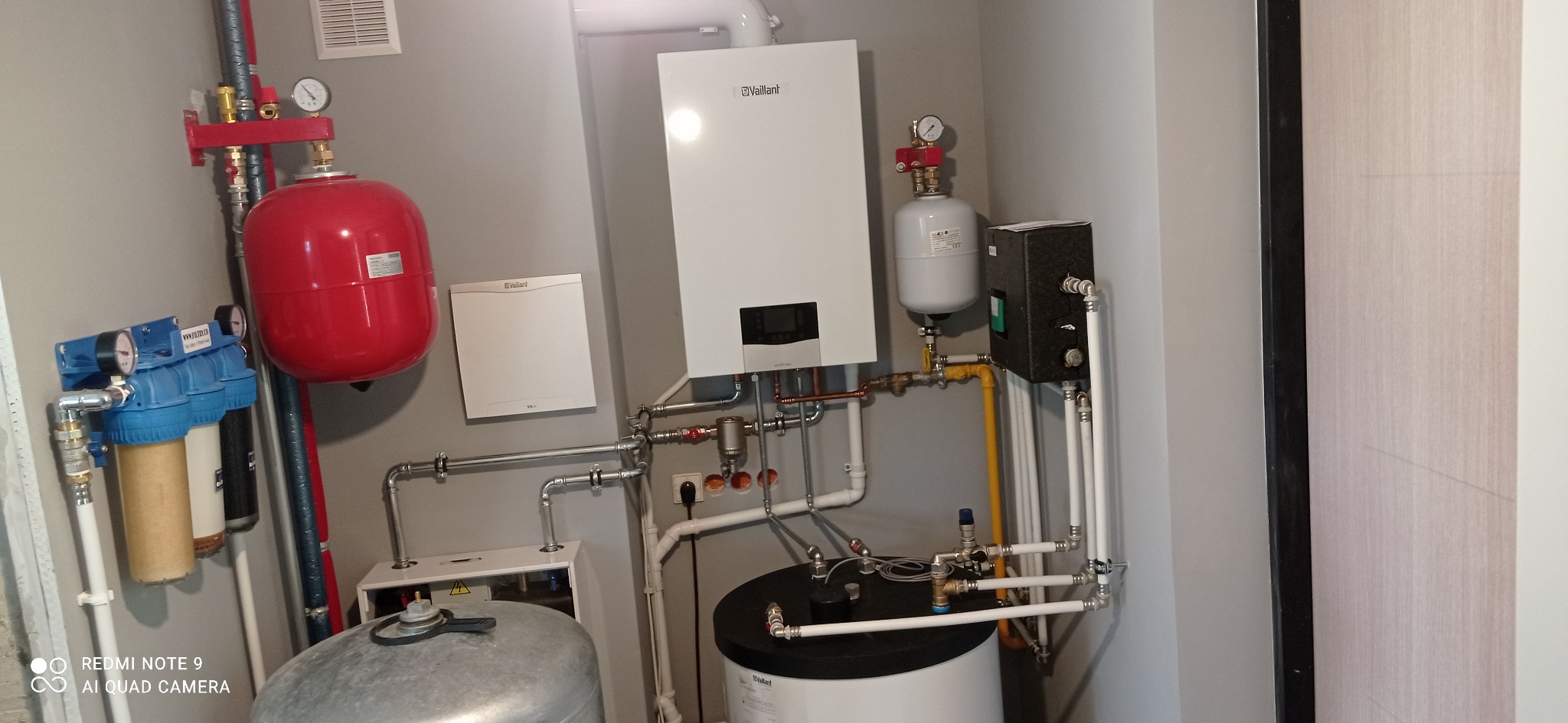 Kompletna kotłownia z piecem gazowym Vaillant, zbiornikiem buforowym, naczyniami przeponowymi i filtrami wody, instalacja w szarym pomieszczeniu z wentylacją.