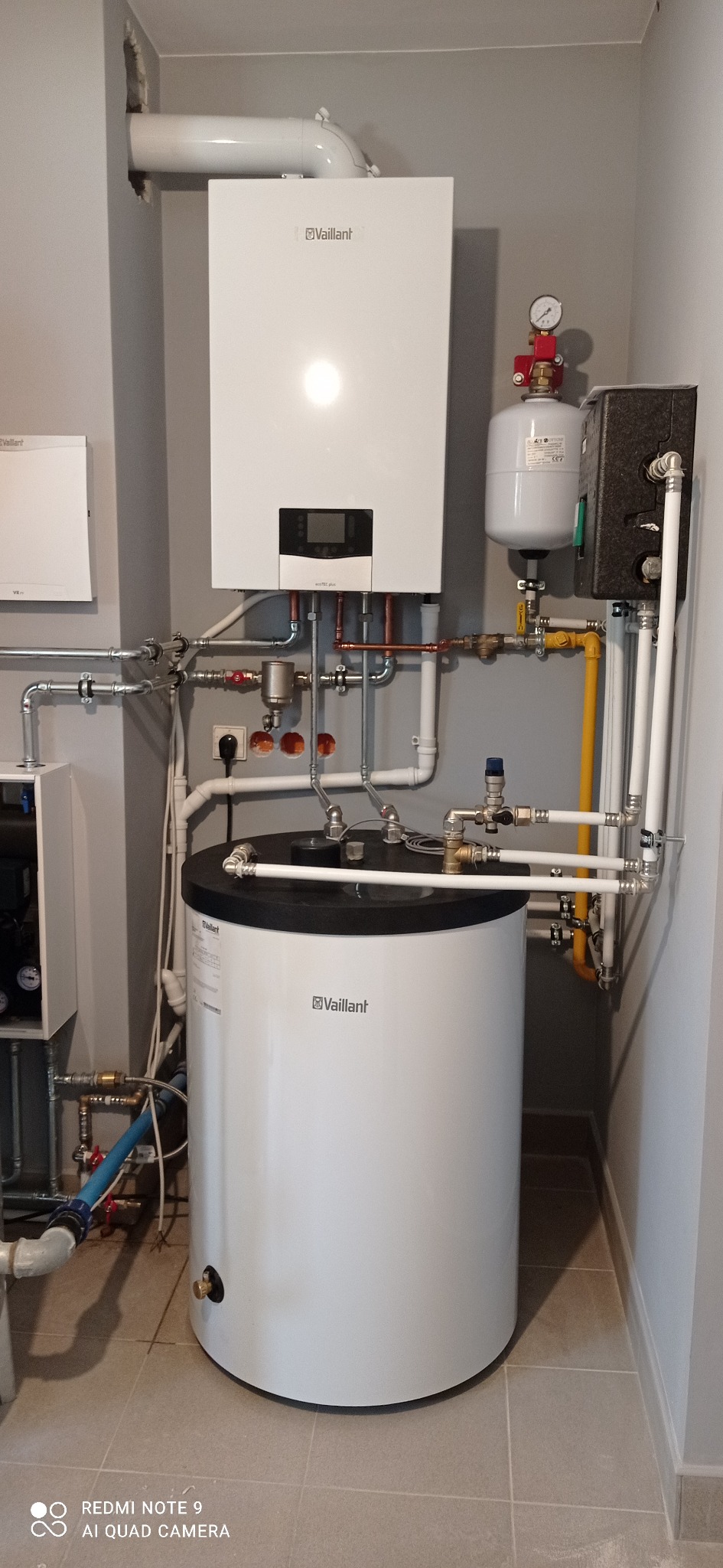 Biały piec gazowy Vaillant z podłączonym zasobnikiem ciepłej wody, widoczne rury miedziane i stalowe, manometr, instalacja w szarym pomieszczeniu z płytkami podłogowymi.