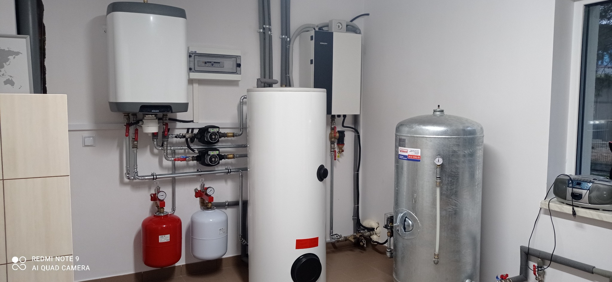 Zestaw urządzeń grzewczych w kotłowni: zbiorniki, pompy obiegowe, naczynia wzbiorcze, instalacja rurowa, szafka elektryczna, radio na parapecie.