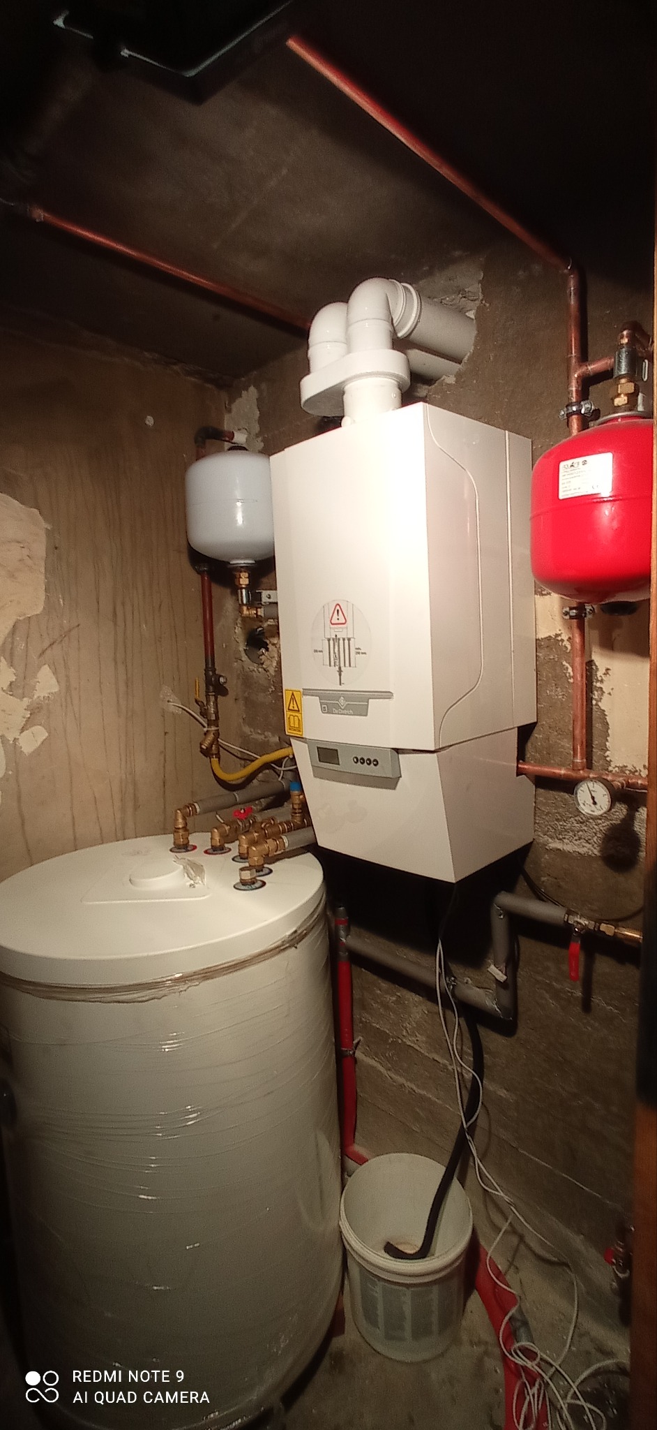 Nowoczesny piec gazowy marki De Dietrich zainstalowany w pomieszczeniu z surowymi ścianami, obok biały zasobnik ciepłej wody i czerwony zbiornik wyrównawczy, widoczne miedziane rury i białe rury...