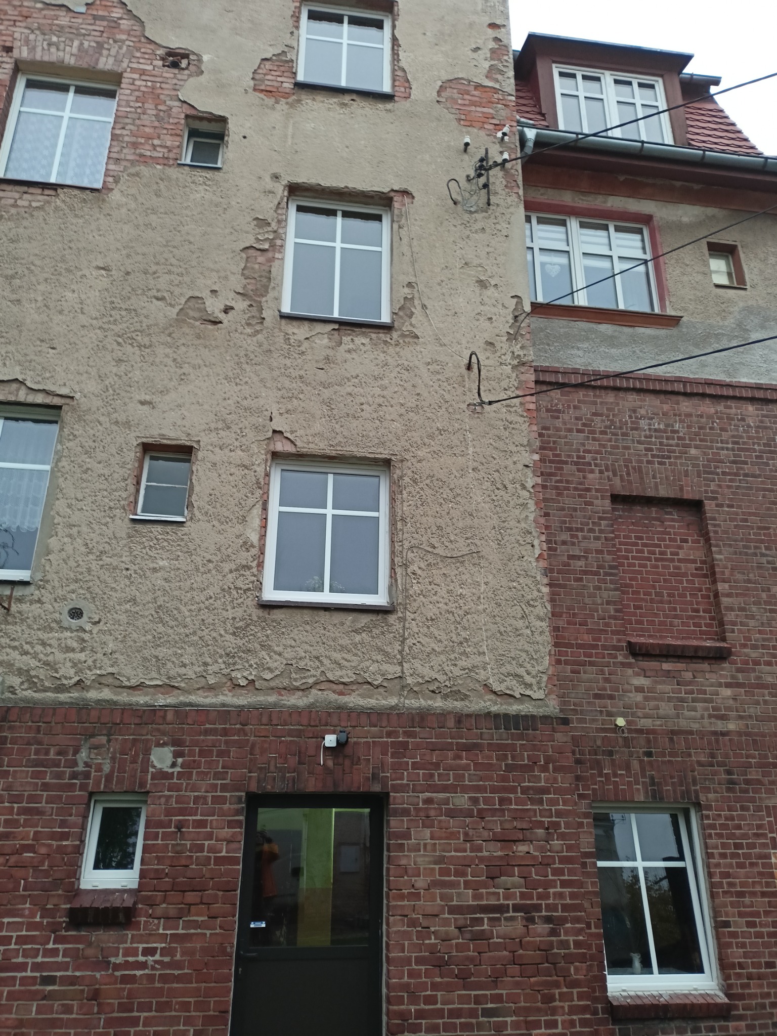 Fasada budynku z mieszanką tynkowanej i ceglanej elewacji, widoczne okna o różnej wielkości i kształcie, ślady renowacji i napraw.