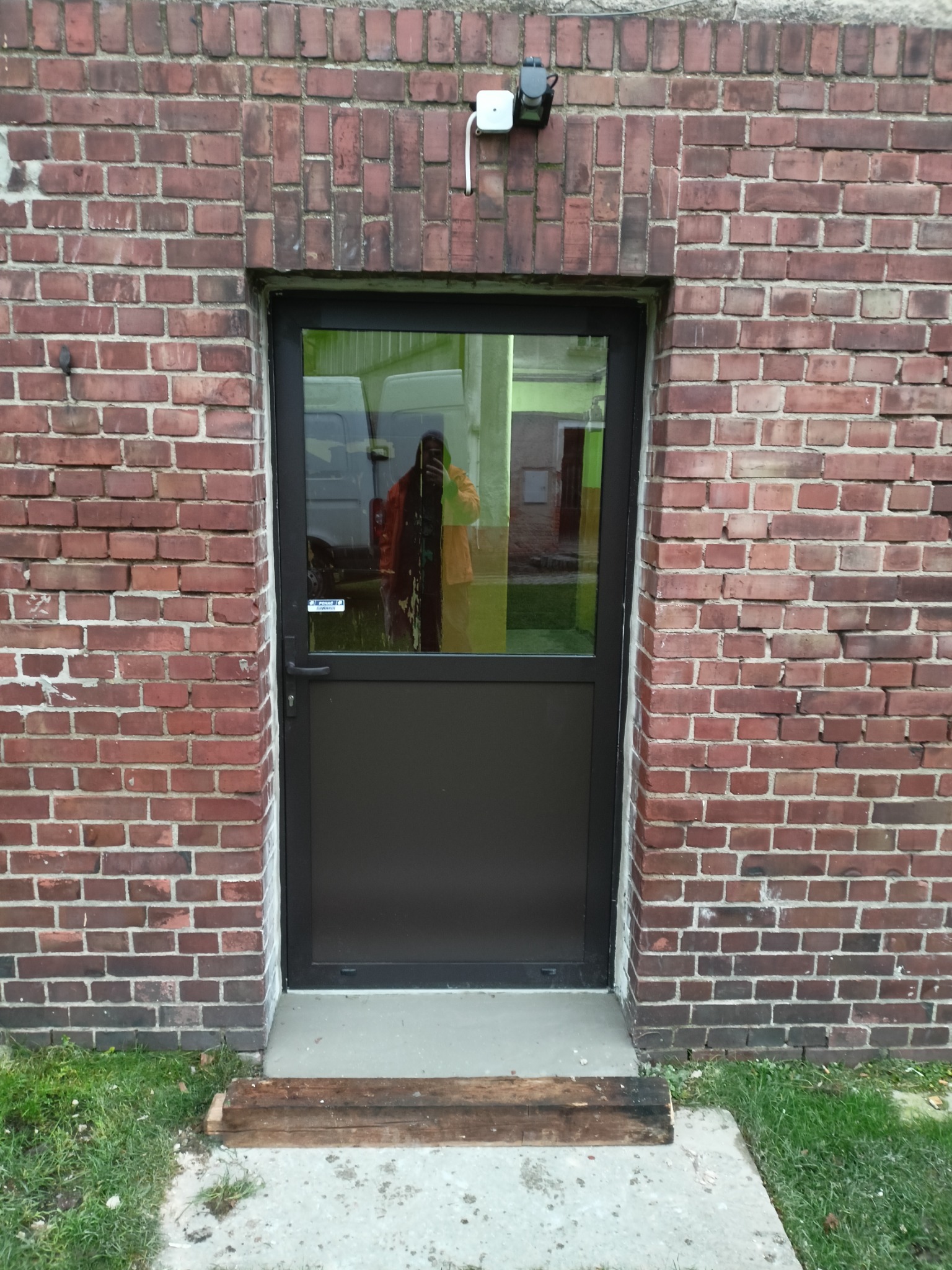 Metalowa rama drzwiowa wmurowana w ścianę z czerwonej cegły, widoczny fragment trawnika i odblask osoby w szybie.