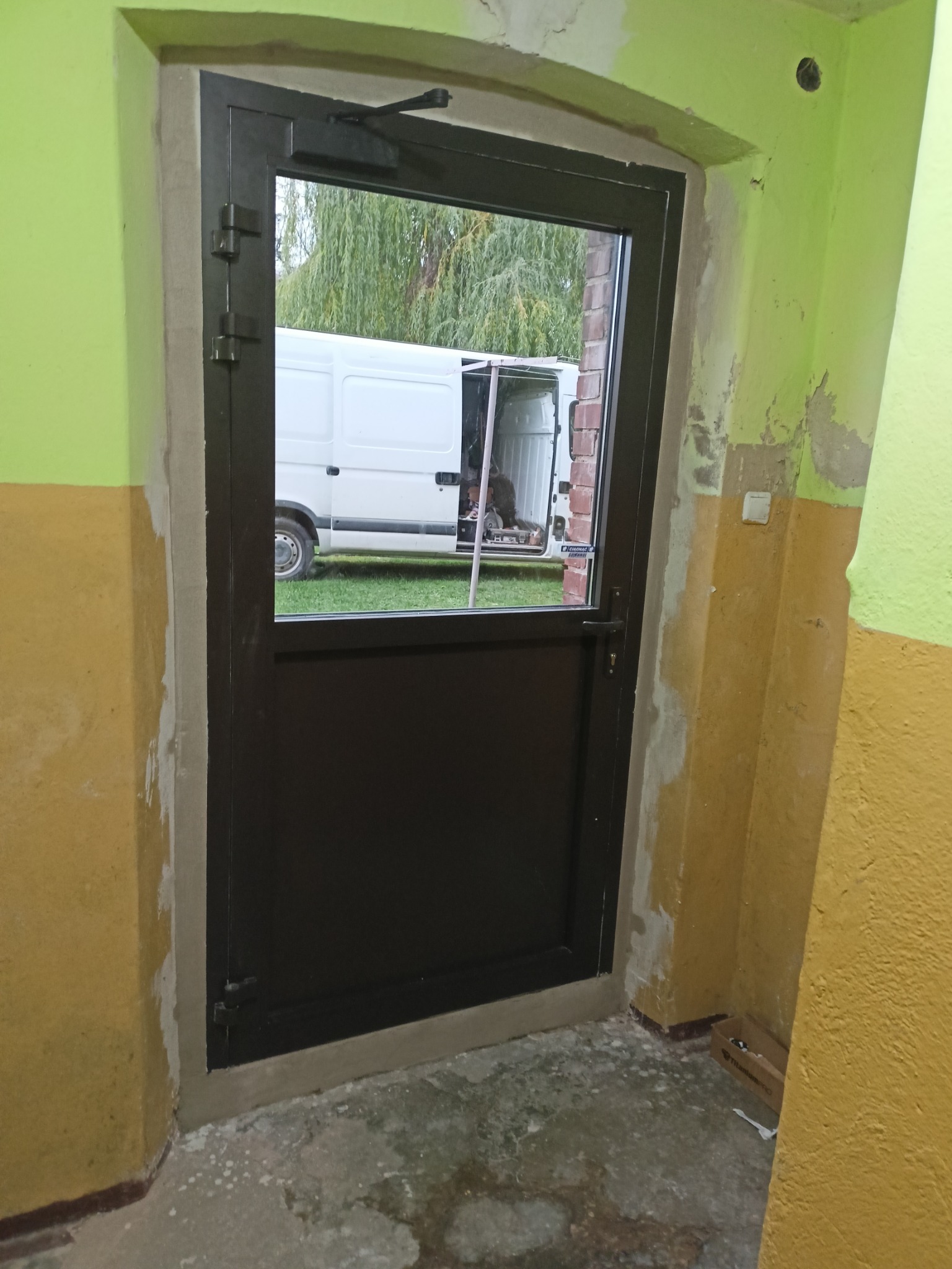 Zamknięte, stalowe drzwi z szybą i samozamykaczem, osadzone w ościeżnicy w surowym stanie, z widocznym na zewnątrz białym busem na trawie.