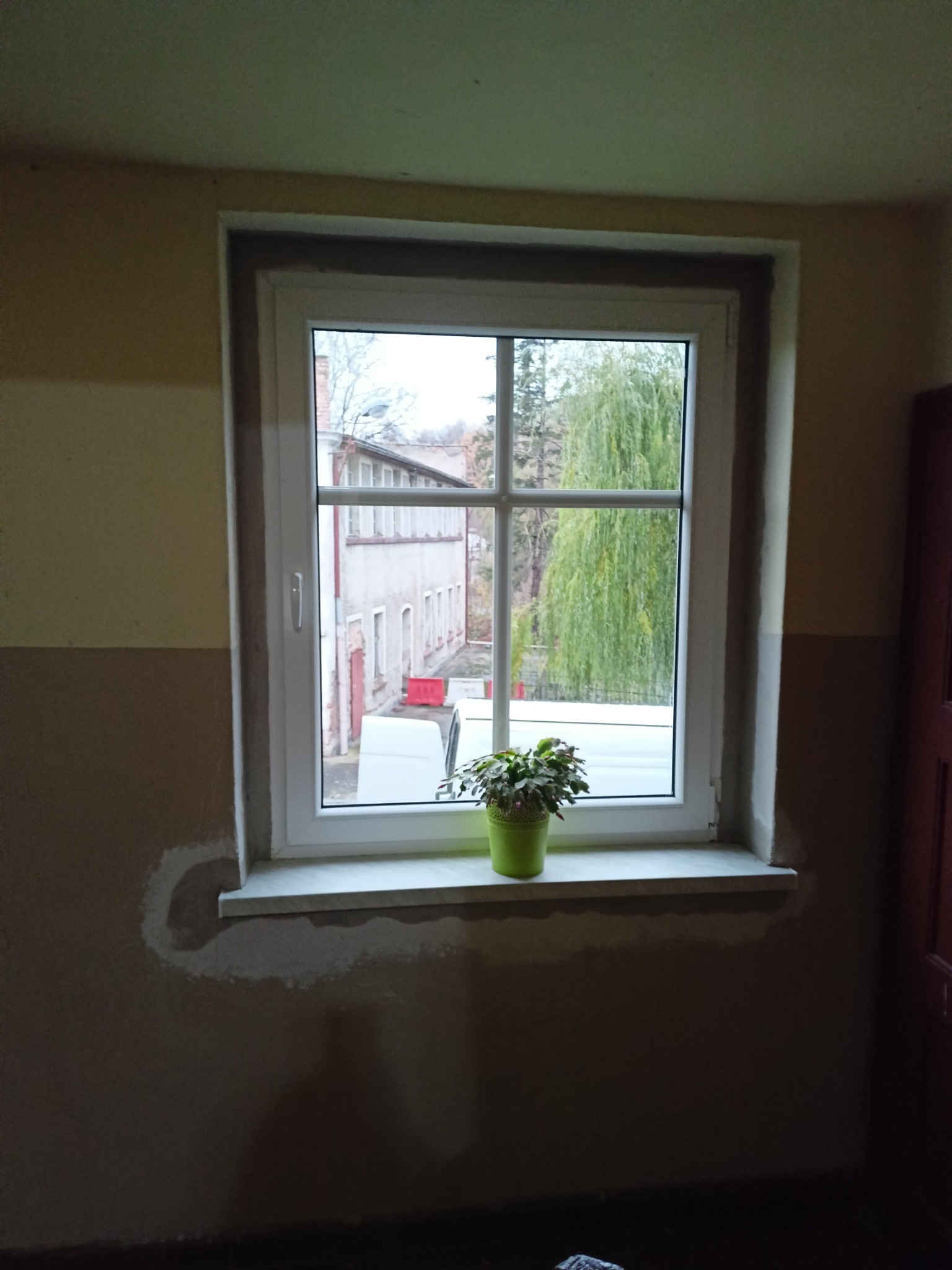 Białe okno PCV z parapetem, na którym stoi zielona doniczka z rośliną, widok na budynek i drzewo zza okna, ślady poprawek tynku na ścianie.