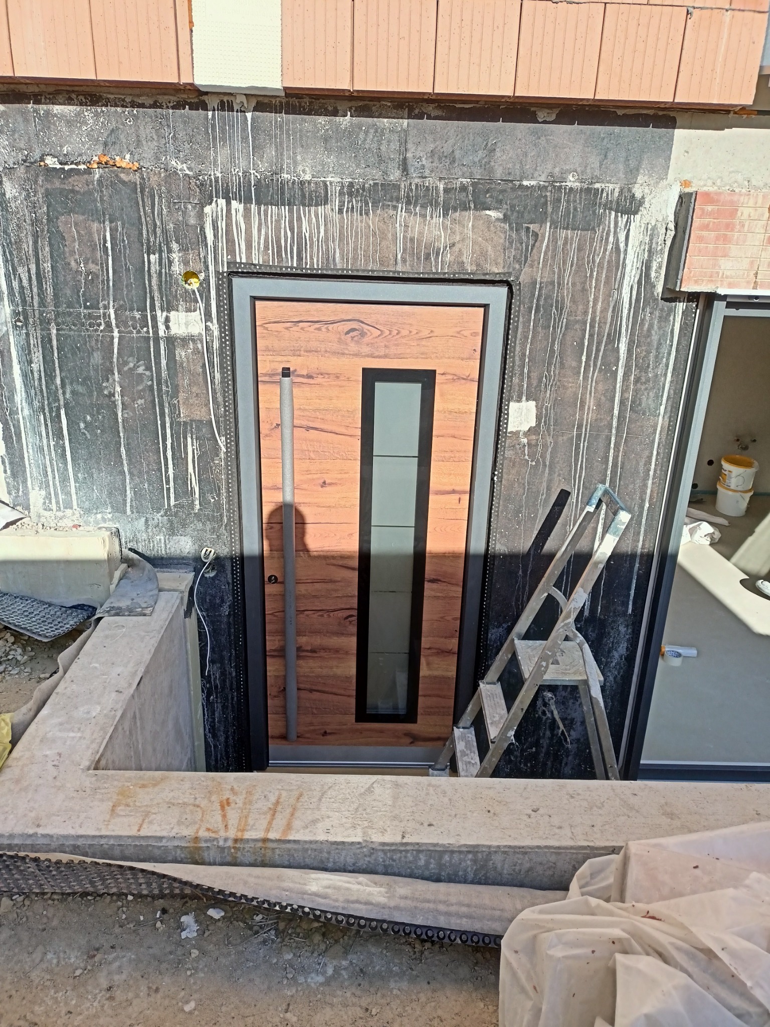 Nowo zamontowane drzwi zewnętrzne z drewnopodobnym panelem i pionowym przeszkleniem, widoczne w surowym otoczeniu budowy z zaciekami na ścianie i drabiną opartą o framugę.