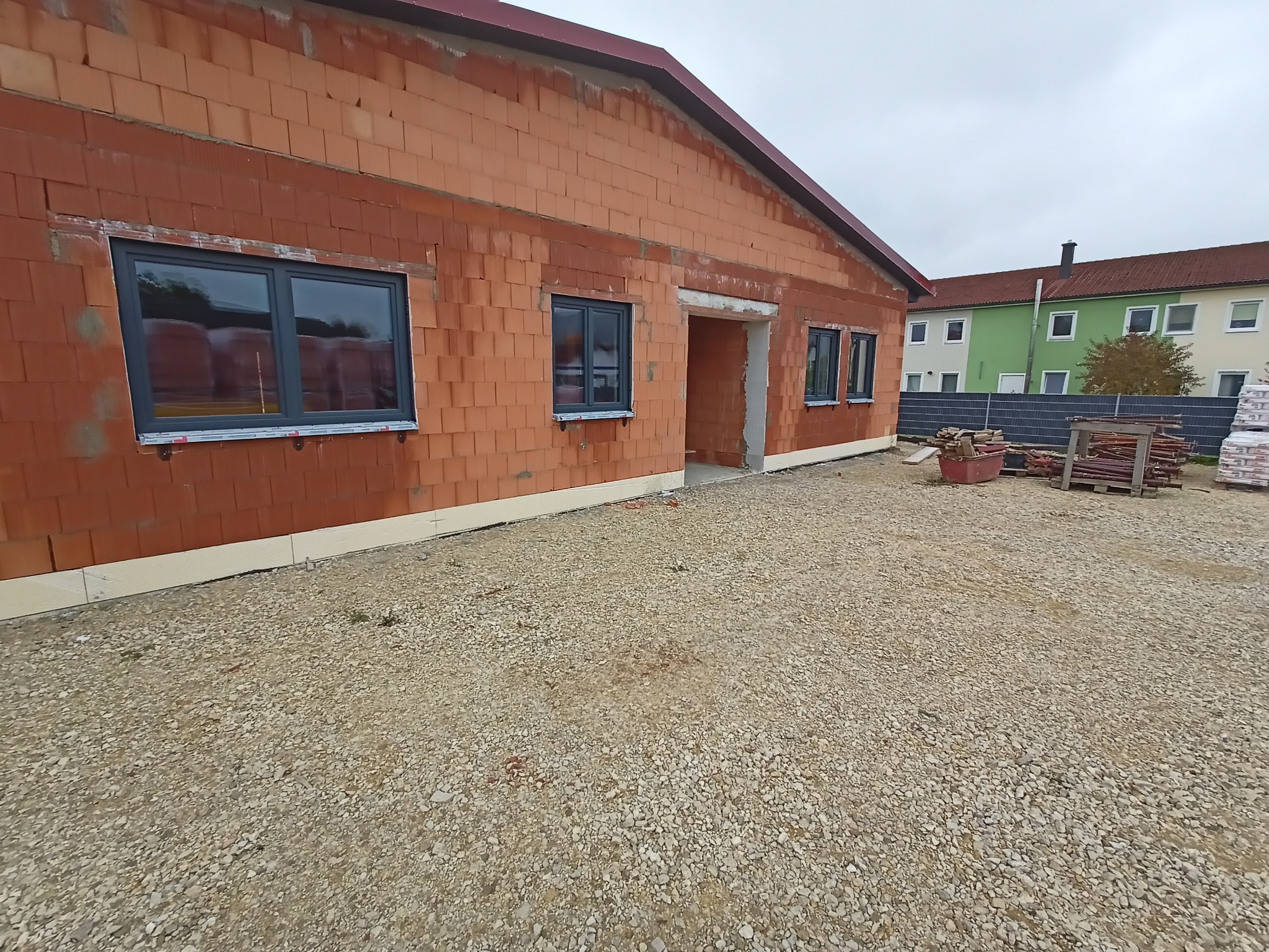 Surowy budynek z cegły ceramicznej z zamontowanymi szarymi oknami, widok z zewnątrz na żwirowe podłoże i materiały budowlane w tle.