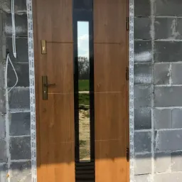 Brązowe drzwi wejściowe z pionowym przeszkleniem, zamontowane w surowym murze z betonu komórkowego, zabezpieczone taśmą montażową i żółtą folią ochronną.