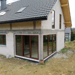 Surowy stan domu jednorodzinnego z zamontowanymi oknami z brązowymi ramami, widoczne bloczki betonowe, szara dachówka i teren budowy z fragmentami gruzu.