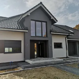 Nowoczesny dom jednorodzinny z szarą elewacją, czarną dachówką, roletami antywłamaniowymi i otwartymi drzwiami wejściowymi, widok z zewnątrz na niezakończone prace brukarskie.