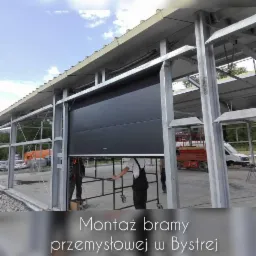 Montaż czarnej bramy segmentowej w metalowej konstrukcji w hali przemysłowej, widoczni monterzy w kombinezonach i podnośnik koszowy w tle.