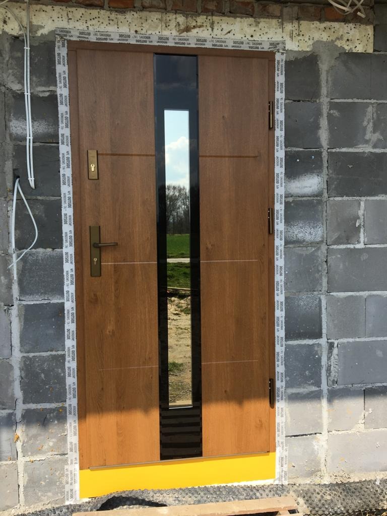 Brązowe drzwi wejściowe z pionowym przeszkleniem, zamontowane w surowym murze z betonu komórkowego, zabezpieczone taśmą montażową i żółtą folią ochronną.