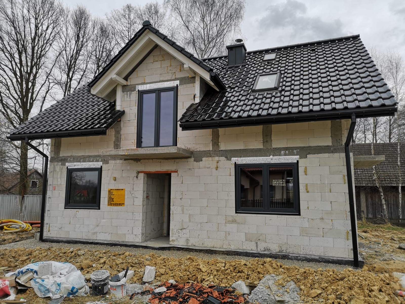 Dom w trakcie budowy z czarnymi ramami okiennymi i ciemną dachówką; widoczne bloczki z betonu komórkowego i niezagospodarowany teren.