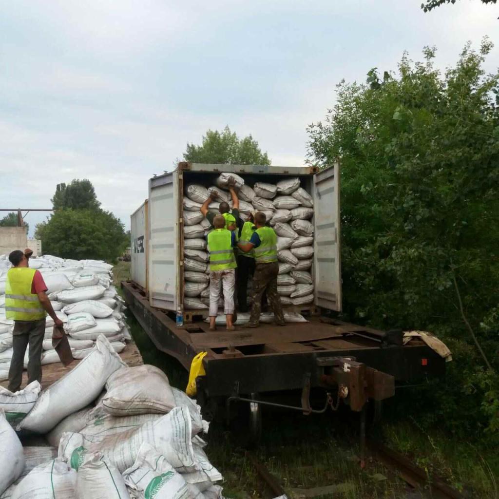 Pracownicy w jaskrawozielonych kamizelkach ładują worki z nawozem do kontenera na platformie kolejowej, widoczne worki na platformie i obok torów, pochmurne niebo w tle.