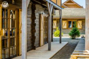 Drewniana zabudowa w stylu western z werandą w Zatorze. Widoczne drzwi z szybami, drewniane ściany i fragment budynku z oknami z okiennicami.