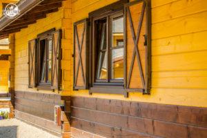 Żółta ściana drewnianego domu z dwoma oknami z brązowymi okiennicami w kształcie litery X. Dolna część ściany pomalowana na brązowo.