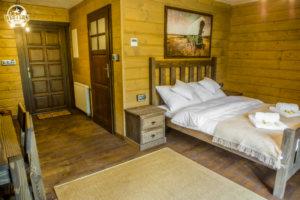 Drewniany pokój gościnny z łóżkiem, szafką nocną i obrazem na ścianie. Widoczny dywanik na podłodze i drzwi wejściowe.