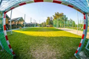 Pusta, zielona murawa boiska sportowego widziana przez siatkę bramki z czerwono-białymi elementami, w tle budynek z drewnianą elewacją i zieleń drzew przy słonecznej pogodzie.