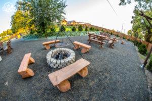 Miejsce na ognisko z kamiennym obramowaniem i drewnianymi ławkami w plenerze, widok z szerokiego kąta, w tle budynek i zieleń.