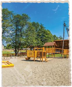 Drewniany plac zabaw z piaskownicą na piaszczystym podłożu, otoczony zielenią drzew i stawem w tle, błękitne niebo