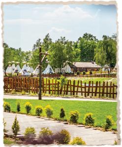 Kompleks rekreacyjny z drewnianym ogrodzeniem, polem namiotowym tipi i budynkiem w tle, widok z perspektywy ścieżki.