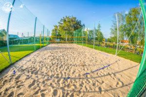 Piaszczyste boisko do siatkówki plażowej z siatką, otoczone zieloną trawą i ogrodzeniem z siatki, w tle drzewa i budynki w słoneczny dzień.