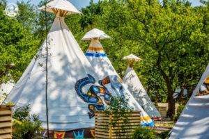 Białe tipi z kolorowymi, etnicznymi wzorami, ustawione w plenerze, otoczone zielenią drzew. Widoczne drewniane konstrukcje u podstawy namiotów.