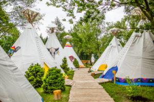 Wioska tipi na imprezę integracyjną: białe namioty z indiańskimi malunkami, drewniana ścieżka, otoczenie zieleni.