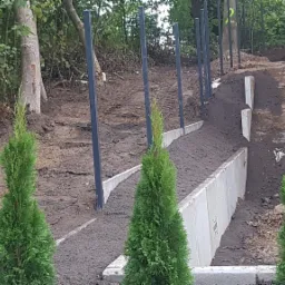 W Sierakowicach budowa betonowego murku oporowego z wbitymi słupkami ogrodzeniowymi i posadzonymi tujami na nierównym terenie.