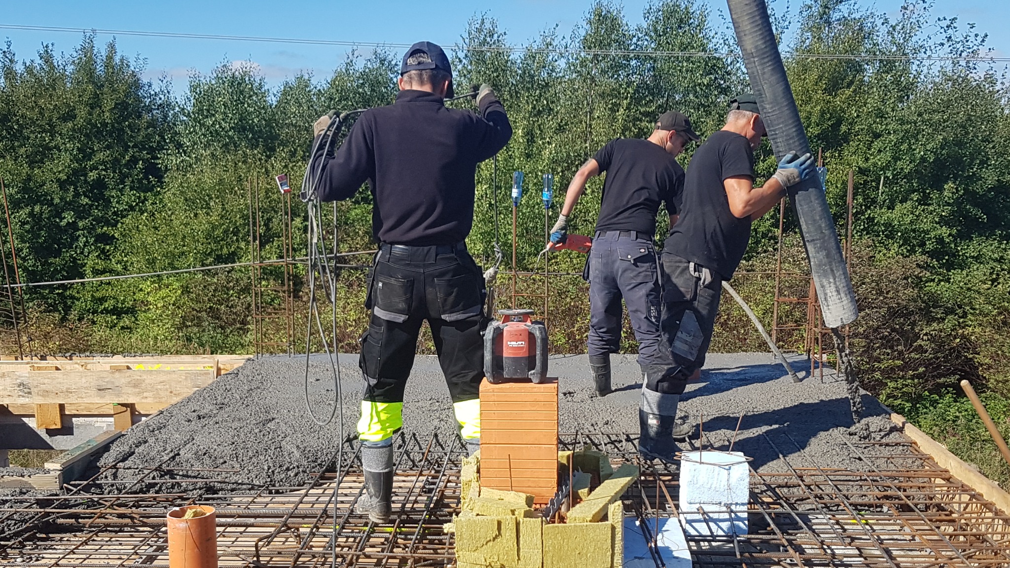 Trzech robotników w czarnych ubraniach wylewa beton na zbrojenie fundamentu. Widoczny laserowy niwelator Hilti na cegłach i izolacji. Tło stanowią zielone drzewa.