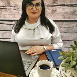 Kobieta w okularach siedzi przy biurku z laptopem, kawą i notatnikiem, prawdopodobnie podczas pracy lub spotkania online.