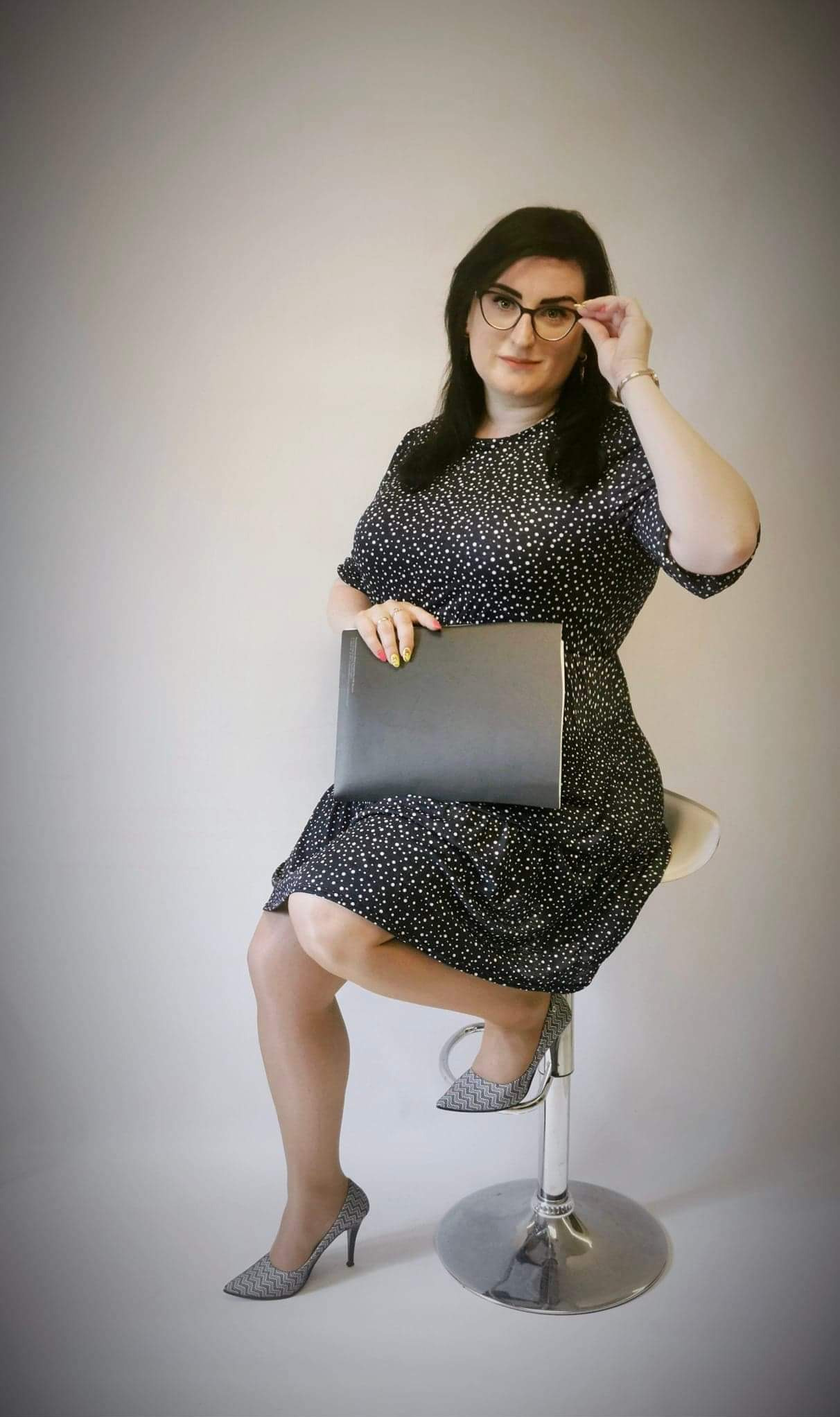 Kobieta w okularach, siedząca na chromowanym stołku barowym, trzyma czarną teczkę, z neonowymi paznokciami i wzorzystych szpilkach, poprawia okulary.