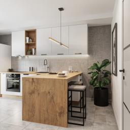 Osky Justyna Knapek - Nowoczesna kuchnia z wyspą, białe szafki wiszące, drewniany blat wyspy z hokerami, betonowa ściana w tle, designerska lampa wisząca, duża roślina doniczkowa obok drzwi.