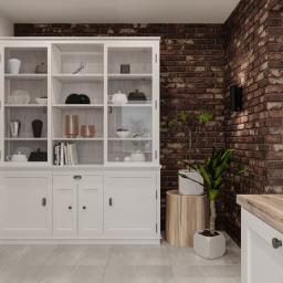Osky Justyna Knapek - Biały kredens z przeszkloną górną częścią, wypełniony dekoracyjnymi naczyniami i książkami, na tle ściany z czerwonej cegły i roślin doniczkowych.
