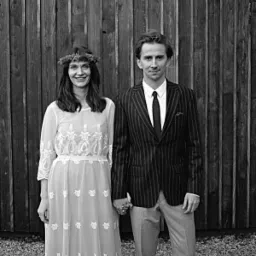 Czarno-białe zdjęcie kobiety w długiej, koronkowej sukni i wianku na głowie, trzymającej za rękę mężczyznę w prążkowanym garniturze na tle drewnianej ściany.