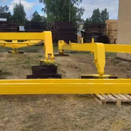 Żółte, pomalowane elementy konstrukcyjne dźwigu leżące na paletach na zewnątrz budynku, z widocznymi śladami odpryskującej farby.