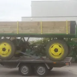 Przyczepa rolnicza z drewnianą skrzynią ładunkową, z żółtymi kołami, transportowana na platformie przyczepki samochodowej, na tle budynku.