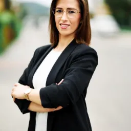 Portret uśmiechniętej kobiety w okularach i czarnej marynarce, z założonymi rękami, na rozmytym tle ulicy.