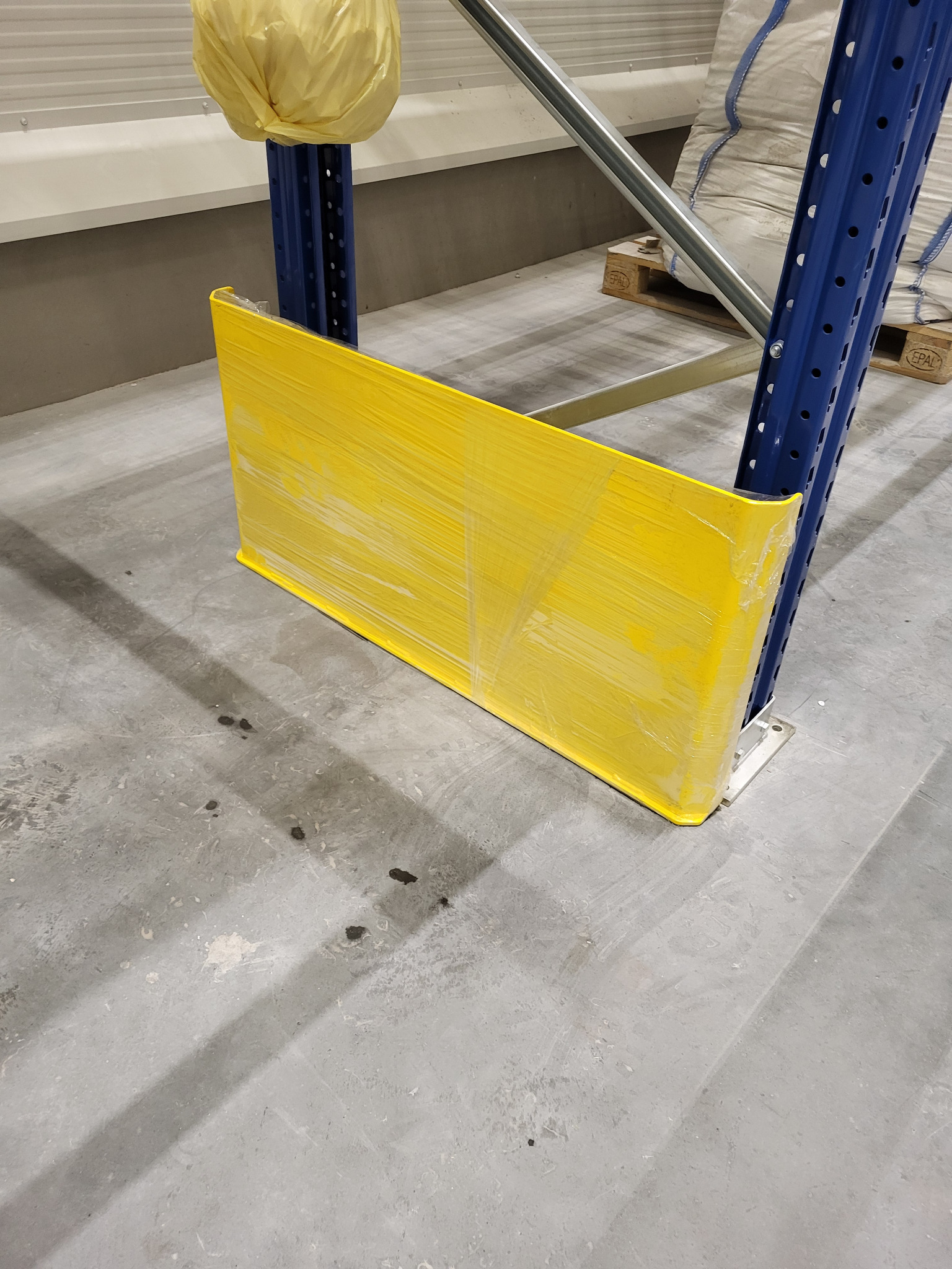 Żółta odbojnica z tworzywa sztucznego, owinięta folią, chroniąca niebieski, metalowy słupek regału magazynowego. Szary beton na podłodze.