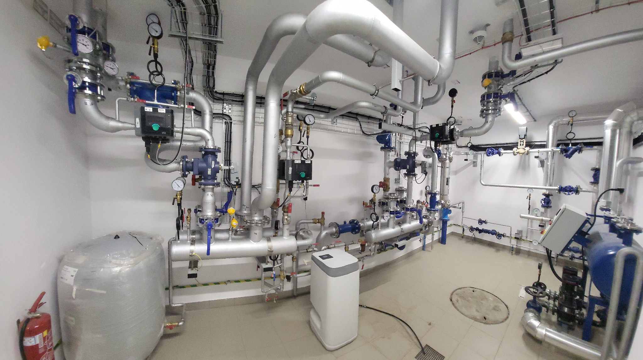 Węzeł cieplny w kotłowni: srebrne rury, zawory, manometry i zbiorniki w czystym, białym pomieszczeniu. Widoczne kable i elementy sterujące.