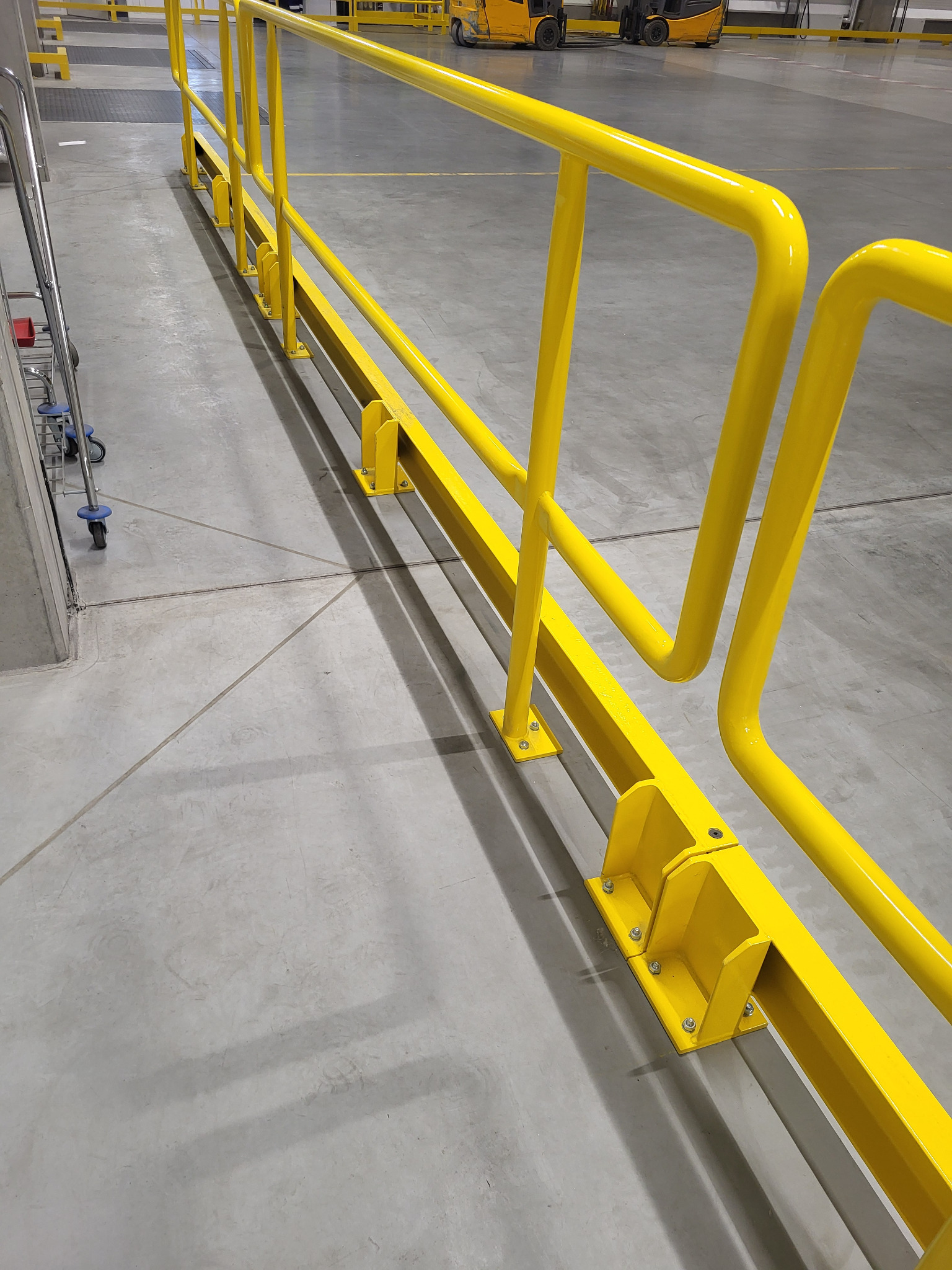 Żółta, metalowa barierka ochronna w hali magazynowej w Kaliszu. Widoczne mocowania do podłoża i fragment wózka transportowego. Jasne, neutralne tło.